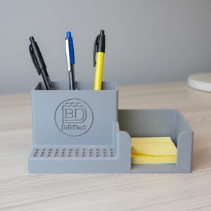 Op de afbeelding: Een grijze bureau-organizer met drie pennen en een stapel gele plaknotities. De organizer heeft een vak voor pennen en een apart gedeelte voor de plaknotities. De organizer heeft het "BrikDock"-logo.