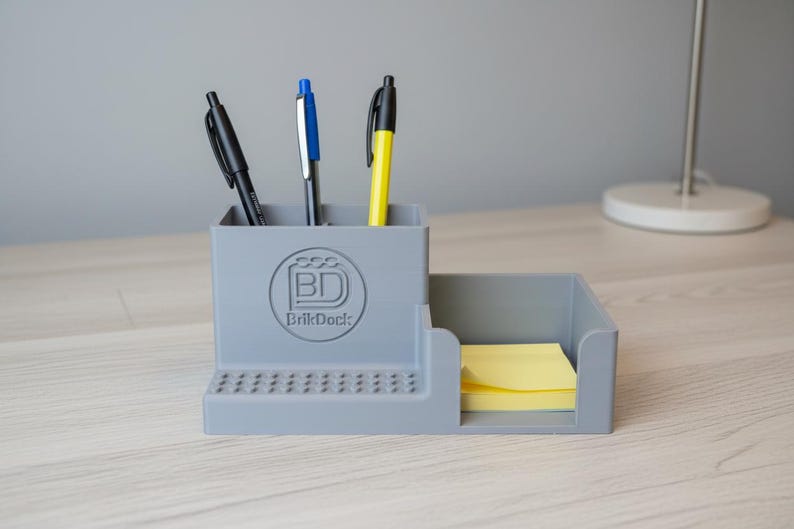 Op de afbeelding: Een grijze bureau-organizer met drie pennen en een stapel gele plaknotities. De organizer heeft een vak voor pennen en een apart gedeelte voor de plaknotities. De organizer heeft het "BrikDock"-logo.