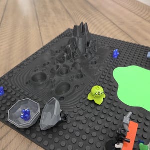 May include: A black Lego baseplate with a 3D-printed lunar landscape, a green blob, and various Lego figures. Includes a gray rock formation, a green alien figure, and blue crystal-like pieces.