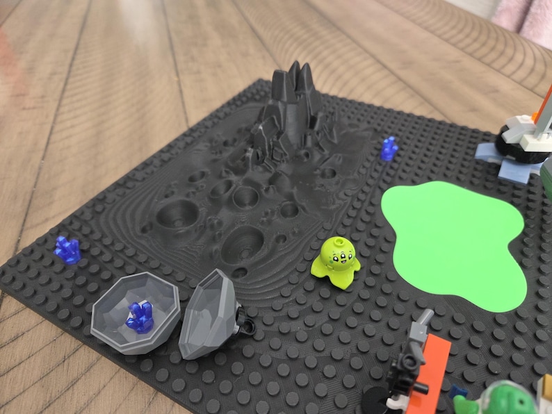 May include: A black Lego baseplate with a 3D-printed lunar landscape, a green blob, and various Lego figures. Includes a gray rock formation, a green alien figure, and blue crystal-like pieces.
