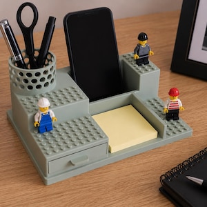 Pode incluir: Um organizador de secret&aacute;ria verde-s&aacute;lvia com um porta-canetas cil&iacute;ndrico perfurado, uma ranhura central para smartphone, uma pequena gaveta e uma superf&iacute;cie plana para notas autocolantes. Tr&ecirc;s pequenas figuras de pl&aacute;stico est&atilde;o fixas.