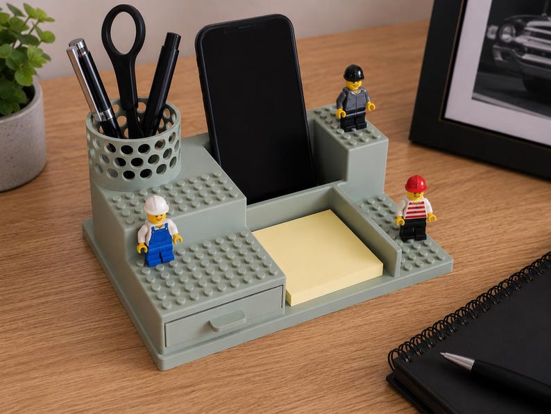 Pode incluir: Um organizador de secret&aacute;ria verde-s&aacute;lvia com um porta-canetas cil&iacute;ndrico perfurado, uma ranhura central para smartphone, uma pequena gaveta e uma superf&iacute;cie plana para notas autocolantes. Tr&ecirc;s pequenas figuras de pl&aacute;stico est&atilde;o fixas.