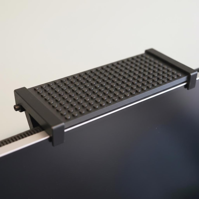 May include: A black, rectangular webcam cover with a textured surface of small, raised dots. The cover is mounted on a silver bar and attached to a black screen. The design is simple and functional.