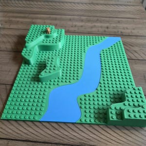 May include: A green plastic building block baseplate with a blue river and green raised terrain. A small brown figurine is on the raised terrain. The baseplate has many small studs.