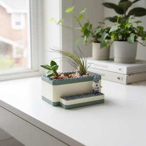 May include: A rectangular planter made from white and green blocks, holding small plants and pebbles. A miniature figure stands on a step-like structure. Other potted plants are in the background, near a window.