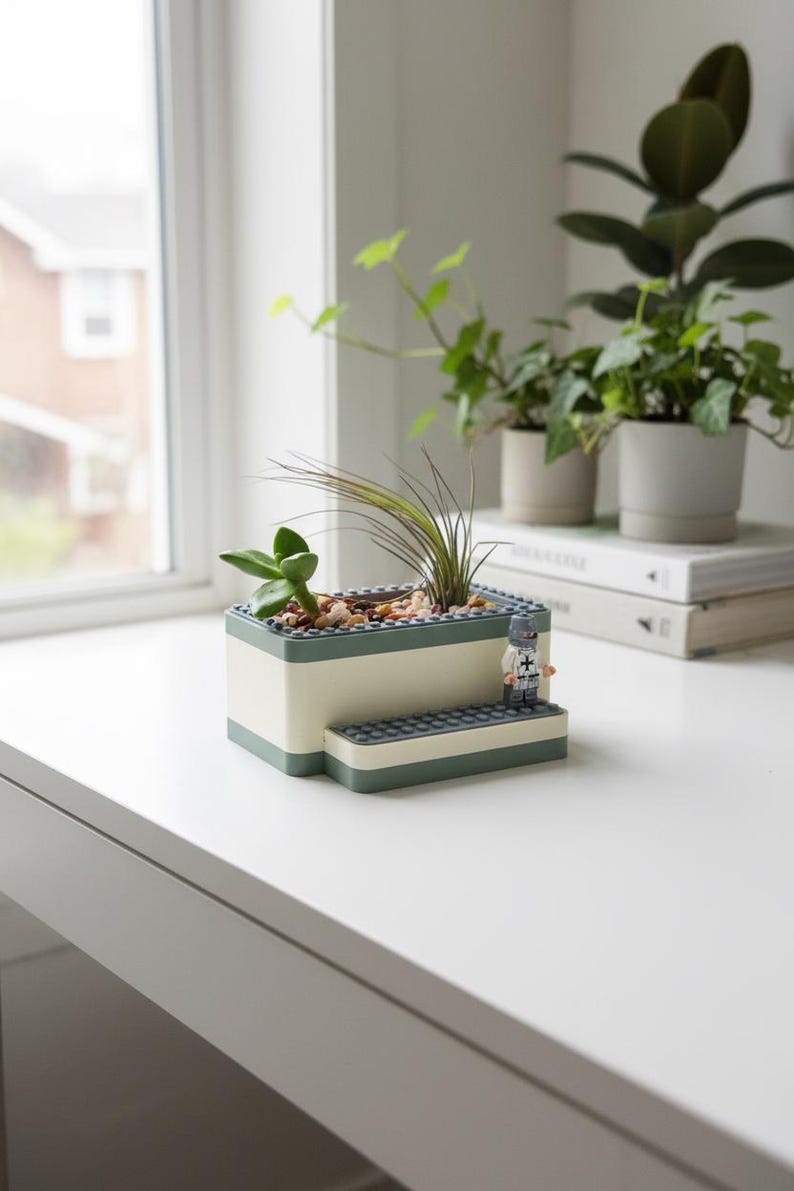 May include: A rectangular planter made from white and green blocks, holding small plants and pebbles. A miniature figure stands on a step-like structure. Other potted plants are in the background, near a window.