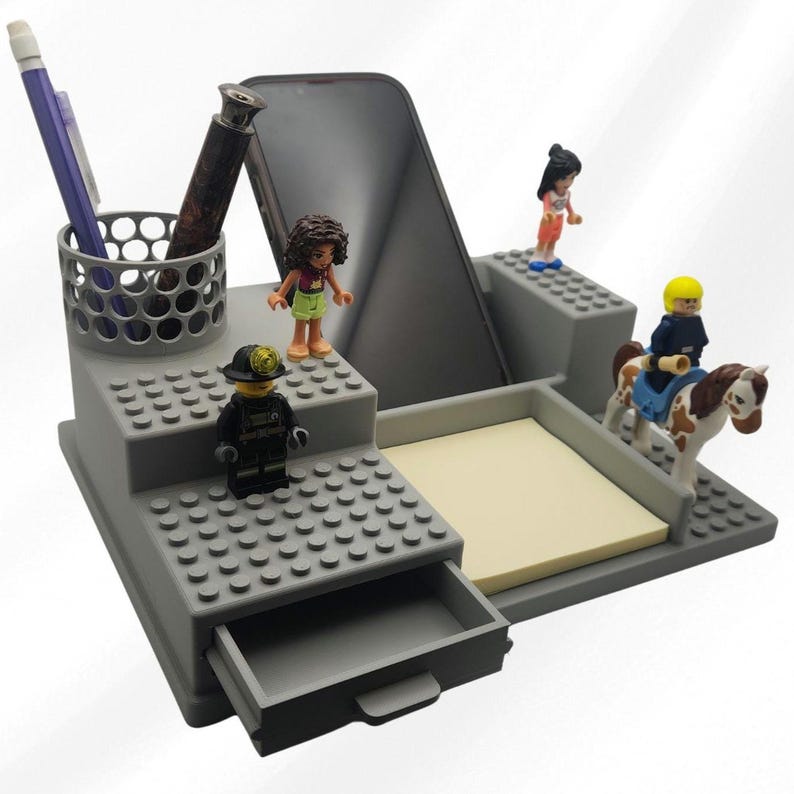 Pode incluir: Um organizador de secret&aacute;ria cinzento com um suporte cil&iacute;ndrico para canetas, um suporte para smartphone, uma pequena gaveta e uma superf&iacute;cie plana para notas autocolantes. O organizador est&aacute; decorado com quatro pequenas figuras de pl&aacute;stico.