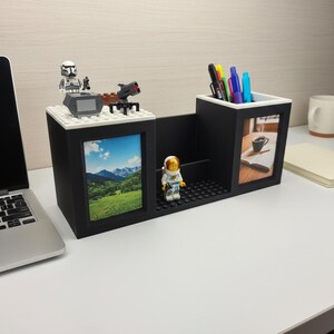 May include: A black desk organizer with a white trim, featuring a picture frame, pen holder, and a small shelf. The organizer is decorated with Lego figures and a landscape photo. Pens and pencils are visible in the pen holder.