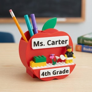 May include: A red apple-shaped desk organizer featuring a green leaf, a yellow pencil, a teal marker, and a purple marker. The front displays white rectangular signs reading Ms. Carter and 4th Grade, with a small plastic figure and building blocks.