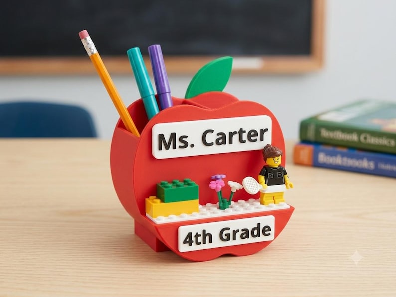 May include: A red apple-shaped desk organizer featuring a green leaf, a yellow pencil, a teal marker, and a purple marker. The front displays white rectangular signs reading Ms. Carter and 4th Grade, with a small plastic figure and building blocks.