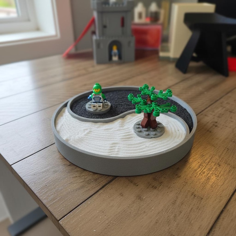 May include: A grey, round miniature zen garden with a yin-yang design. The garden features white sand, black gravel, and Lego figures of a tree and a person. The zen garden is on a wooden surface.