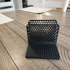 May include: Black phone stand with a honeycomb design. The stand has a rectangular base with small, rounded bumps. The back of the stand features hexagonal cutouts. The stand is made of a matte black material.