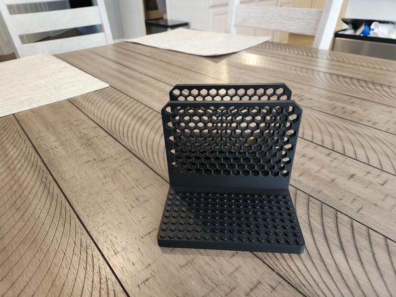 May include: Black phone stand with a honeycomb design. The stand has a rectangular base with small, rounded bumps. The back of the stand features hexagonal cutouts. The stand is made of a matte black material.