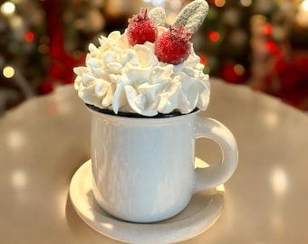 May include: A white ceramic mug with a black rim, filled with whipped cream and topped with two red cherries and two sugar-dusted leaf-shaped decorations. The mug sits on a matching saucer, creating a festive, holiday-themed display.