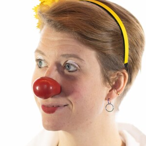 Op de afbeelding: Een persoon met een felrode clownsneus, een gele haarband en een gele bloem. De persoon heeft kort bruin haar, witte make-up en rode lippenstift. De achtergrond is wit.