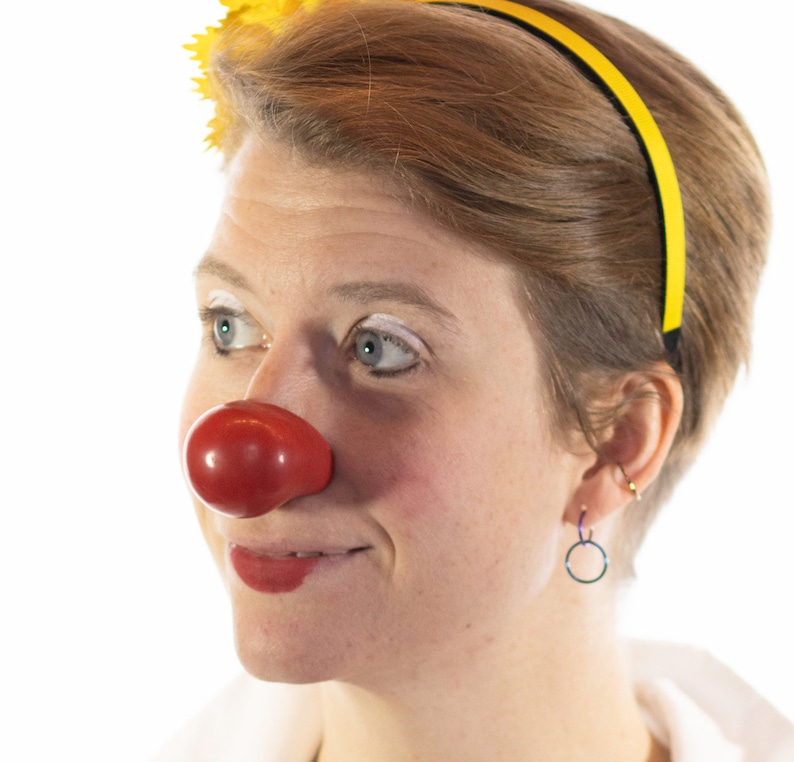 Op de afbeelding: Een persoon met een felrode clownsneus, een gele haarband en een gele bloem. De persoon heeft kort bruin haar, witte make-up en rode lippenstift. De achtergrond is wit.
