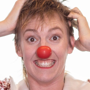 Op de afbeelding: Een persoon met kort, lichtbruin haar, met een rode clownsneus en een wit overhemd met kraag en een bloemenpatroon. De persoon glimlacht breed, met de handen op het hoofd.