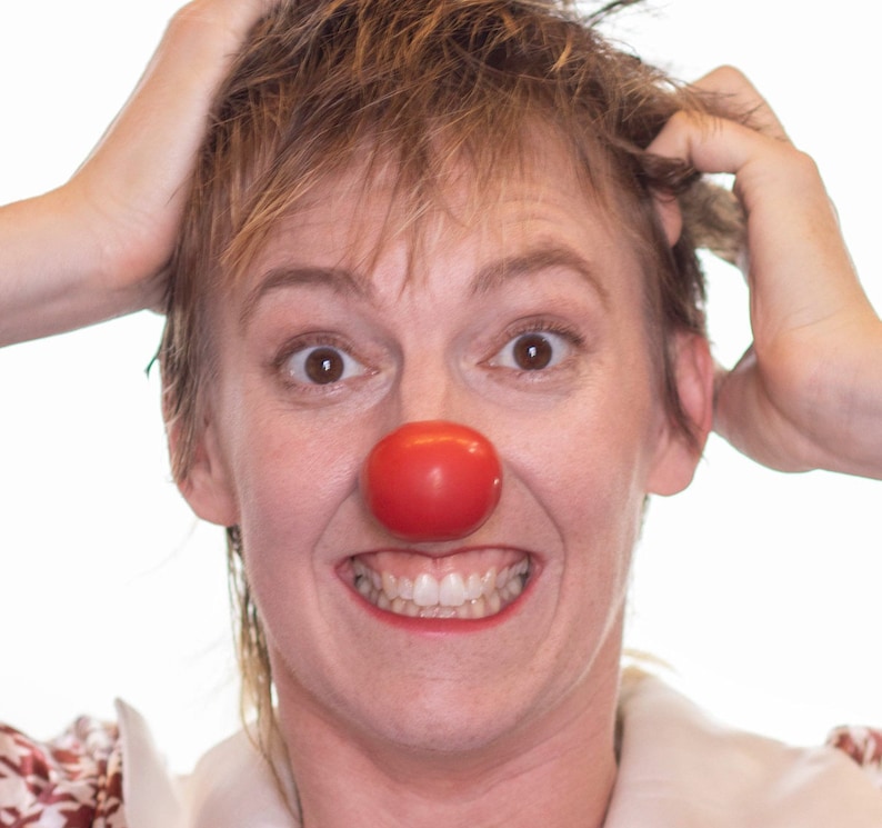 Op de afbeelding: Een persoon met kort, lichtbruin haar, met een rode clownsneus en een wit overhemd met kraag en een bloemenpatroon. De persoon glimlacht breed, met de handen op het hoofd.