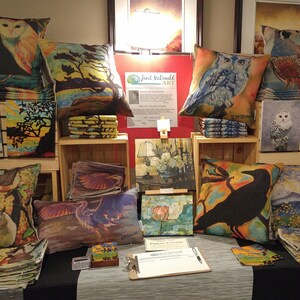 May include: A table display of handmade pillows and artwork featuring nature-inspired designs. The pillows are made with colorful fabrics and feature images of owls, foxes, birds, and other animals. The artwork is also nature-themed and includes paintings of flowers, birds, and landscapes. The display is set up in a room with a light grey carpet and a wooden floor.
