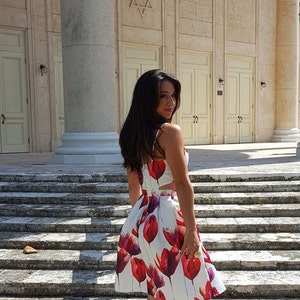 以下が含まれることがあります： 白い花柄の赤いドレスと肌色のヒールを履いた女性が、白い柱のある建物の前の階段に立っています。