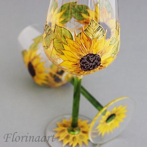 May include: Two clear glass wine glasses with green stems and bases. Each glass is hand-painted with a sunflower design. The sunflowers are yellow with brown centres and green leaves.