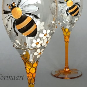 May include: Two hand-painted wine glasses featuring a bee and honeycomb design. The glasses are clear with a gold and black bee painted on the front. The stem of the glasses is painted with a honeycomb pattern.