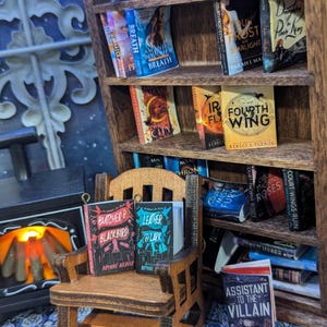 May include: A miniature wooden bookcase filled with small books. A tiny rocking chair holds two books, "Butcher & Blackbird" and "Leather & Lark." A fireplace and patterned rug complete the scene. Bookshelves are a popular item for book lovers.