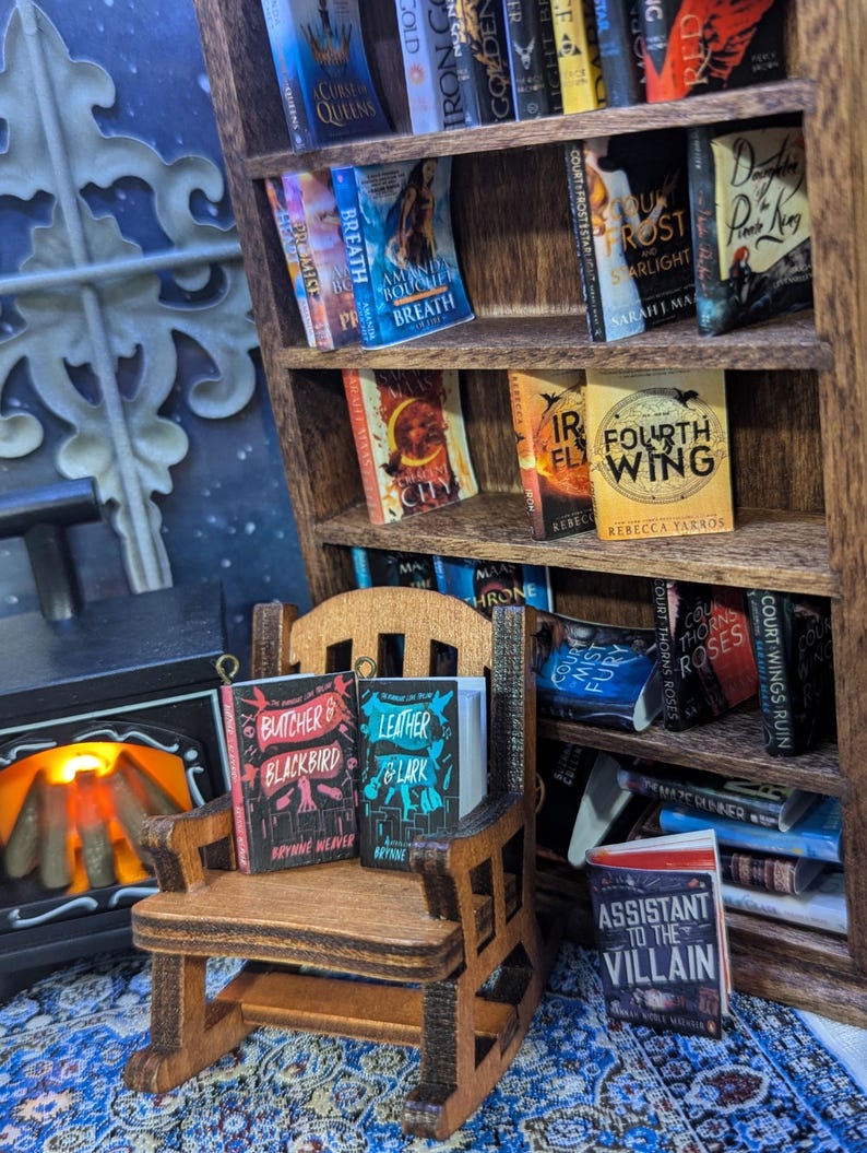 May include: A miniature wooden bookcase filled with small books. A tiny rocking chair holds two books, "Butcher & Blackbird" and "Leather & Lark." A fireplace and patterned rug complete the scene. Bookshelves are a popular item for book lovers.