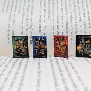 May include: Four miniature books with colourful covers are arranged on a printed page. The books feature titles such as "The Inheritance Games" and "The Final Gambit." The books are part of a series by Jennifer Lynn Barnes.
