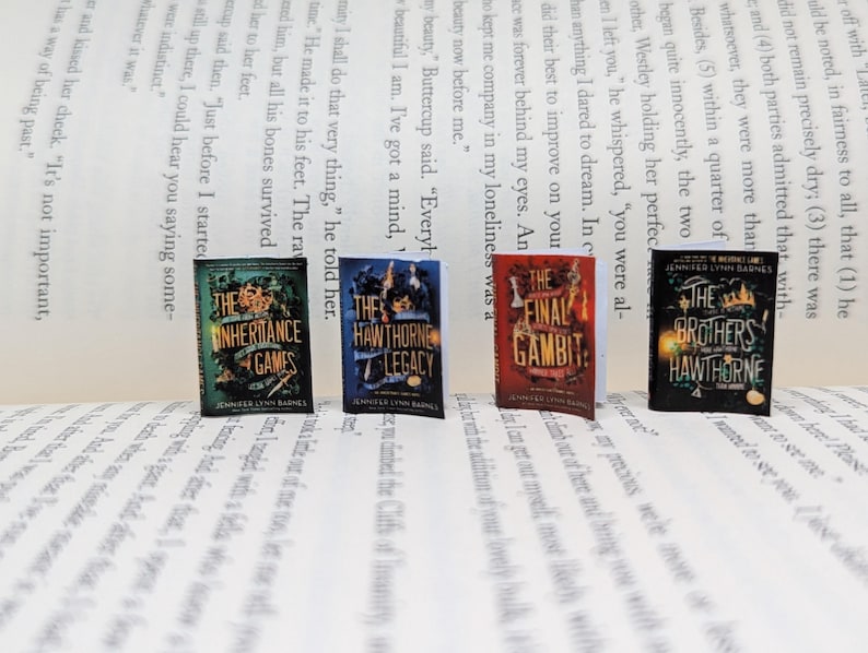 May include: Four miniature books with colourful covers are arranged on a printed page. The books feature titles such as "The Inheritance Games" and "The Final Gambit." The books are part of a series by Jennifer Lynn Barnes.
