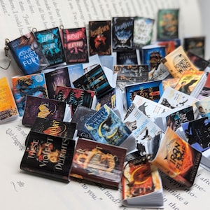 May include: A pile of small, colourful books with fantasy and romance themes, resting on the open pages of a book. The miniature books display titles such as "Court of Mist and Fury" and "Iron Flame", creating a literary display.