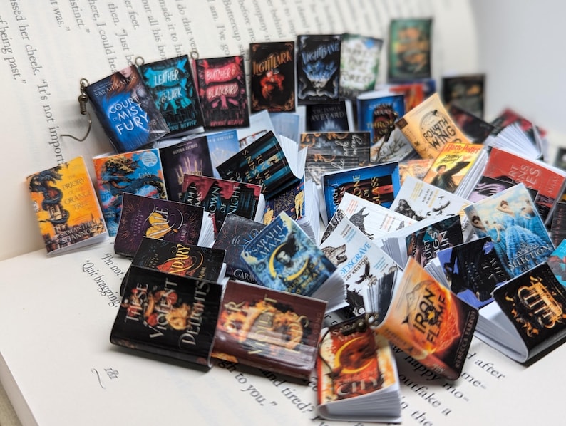 May include: A pile of small, colourful books with fantasy and romance themes, resting on the open pages of a book. The miniature books display titles such as "Court of Mist and Fury" and "Iron Flame", creating a literary display.