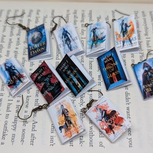 May include: A collection of miniature book earrings, each with a different cover design and title, including "Assassin's Blade" and "Kingdom of Ash." The earrings are displayed on a book page and have bronze-toned hooks. The covers are colourful.