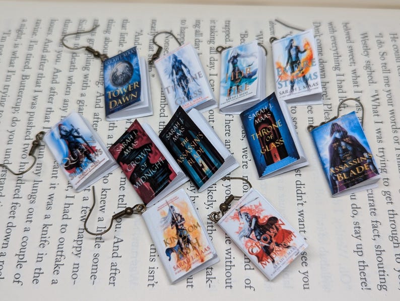 May include: A collection of miniature book earrings, each with a different cover design and title, including "Assassin's Blade" and "Kingdom of Ash." The earrings are displayed on a book page and have bronze-toned hooks. The covers are colourful.
