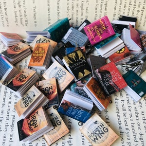 May include: A pile of small, colourful books, including titles such as "Iron Flame" and "Fourth Wing", are arranged on a printed page. Each miniature book has a small metal loop attached to it.