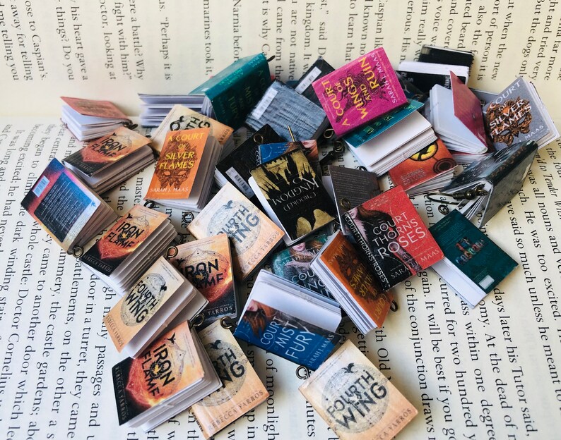 May include: A pile of small, colourful books, including titles such as "Iron Flame" and "Fourth Wing", are arranged on a printed page. Each miniature book has a small metal loop attached to it.
