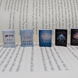 May include: A collection of miniature books, including "Red Queen" and "Glass Sword," displayed on a printed page. The books have vibrant covers with titles and author names. The set features books by Victoria Aveyard.