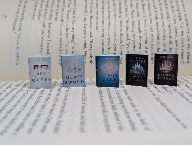 May include: A collection of miniature books, including "Red Queen" and "Glass Sword," displayed on a printed page. The books have vibrant covers with titles and author names. The set features books by Victoria Aveyard.