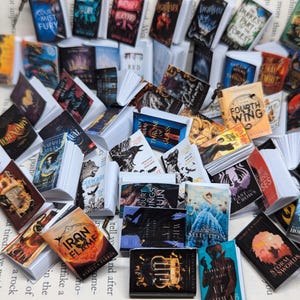 May include: A selection of miniature books with vibrant covers, featuring titles such as "Fourth Wing" and "Iron Flame." The books are displayed on a page of printed text, creating a whimsical, literary scene. The books vary in size and design.
