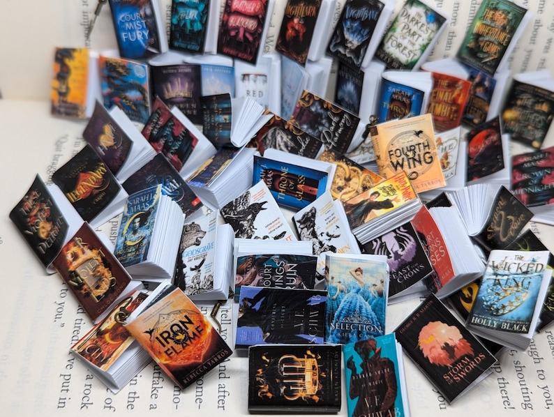May include: A selection of miniature books with vibrant covers, featuring titles such as "Fourth Wing" and "Iron Flame." The books are displayed on a page of printed text, creating a whimsical, literary scene. The books vary in size and design.