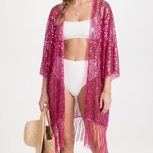 Op de afbeelding: Een felroze kimono-cover-up met lovertjes, een open voorkant, driekwartmouwen en een zoom met lange franjes. Het kledingstuk wordt gedragen over een wit, geribd tweedelig badpak en gecombineerd met een gevlochten rieten tas.