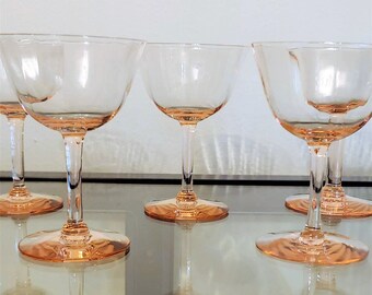 May include: A set of five vintage pink champagne coupe glasses. The glasses have a delicate, rounded bowl and a slender stem, with a matching pink base. The glasses are arranged on a reflective surface, with a white background.