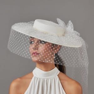 May include: A white hat with a wide brim and a matching veil dotted with small white circles. The hat features a cylindrical crown and a decorative bow. The veil drapes over the face and shoulders, creating an elegant look.
