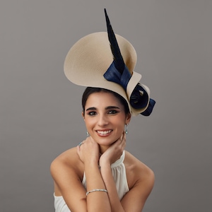 Puede incluir: Un sombrero beige y azul marino con ala ancha y adornos decorativos, incluyendo un gran lazo azul marino y una larga pluma negra. Un accesorio elegante para eventos especiales.