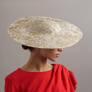 Puede incluir: Un sombrero de ala ancha con un patrón texturizado crema y dorado. El sombrero tiene una copa redondeada y un ala ancha y circular. El sombrero se usa con una blusa roja. El diseño del sombrero sugiere que es adecuado para eventos formales u ocasiones especiales.