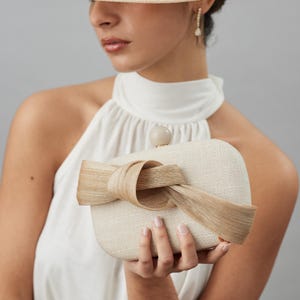 Könnte beinhalten: Eine beigefarbene Clutch mit gewebter Textur und dekorativer Schleife. Die Clutch hat eine abgerundete Form und einen hellfarbigen Verschluss. Ein passender breitkrempiger Hut ist ebenfalls zu sehen. Die Gesamtästhetik ist elegant und für besondere Anlässe geeignet.