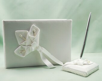 May include: White wedding guest book with a white satin cover and a white ribbon. The book has a white flower design on the front. A white pen holder with a white pen is included.