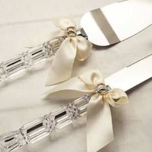 May include: A set of two wedding cake cutting knives with crystal handles and ivory satin bows. Each bow has a gold ring charm.