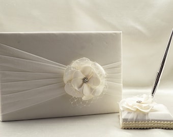 May include: White guest book with a white satin ribbon and a white flower with a rhinestone center. A white pen holder with a white flower and a silver pen.