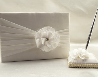 May include: White wedding guest book with a white satin bow and a white flower with a rhinestone center. A matching pen holder with a white flower and a silver pen.